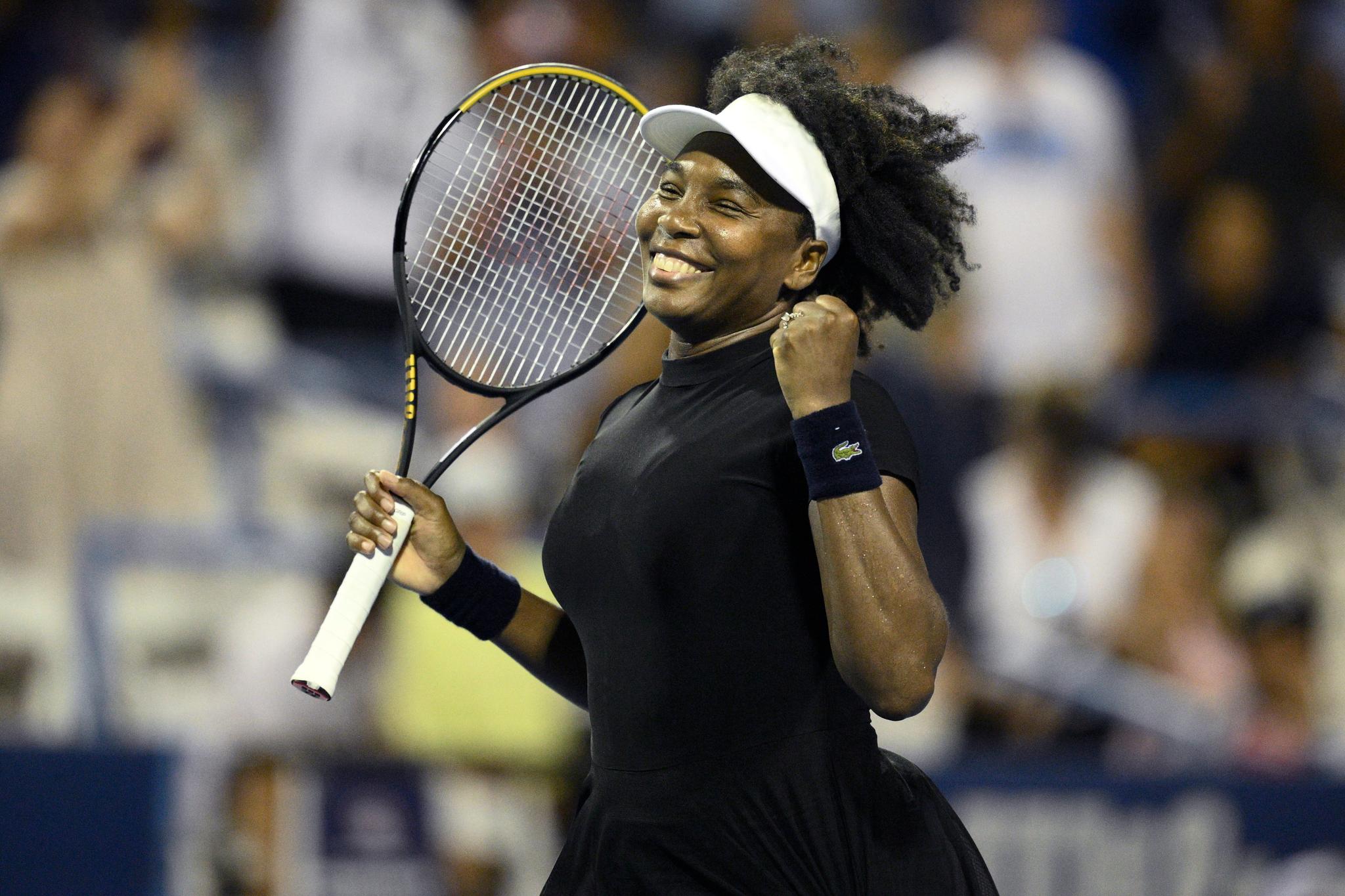Woman, smiling after a win. She's wearing a white visor and holding up a Wilson tennis racquet.