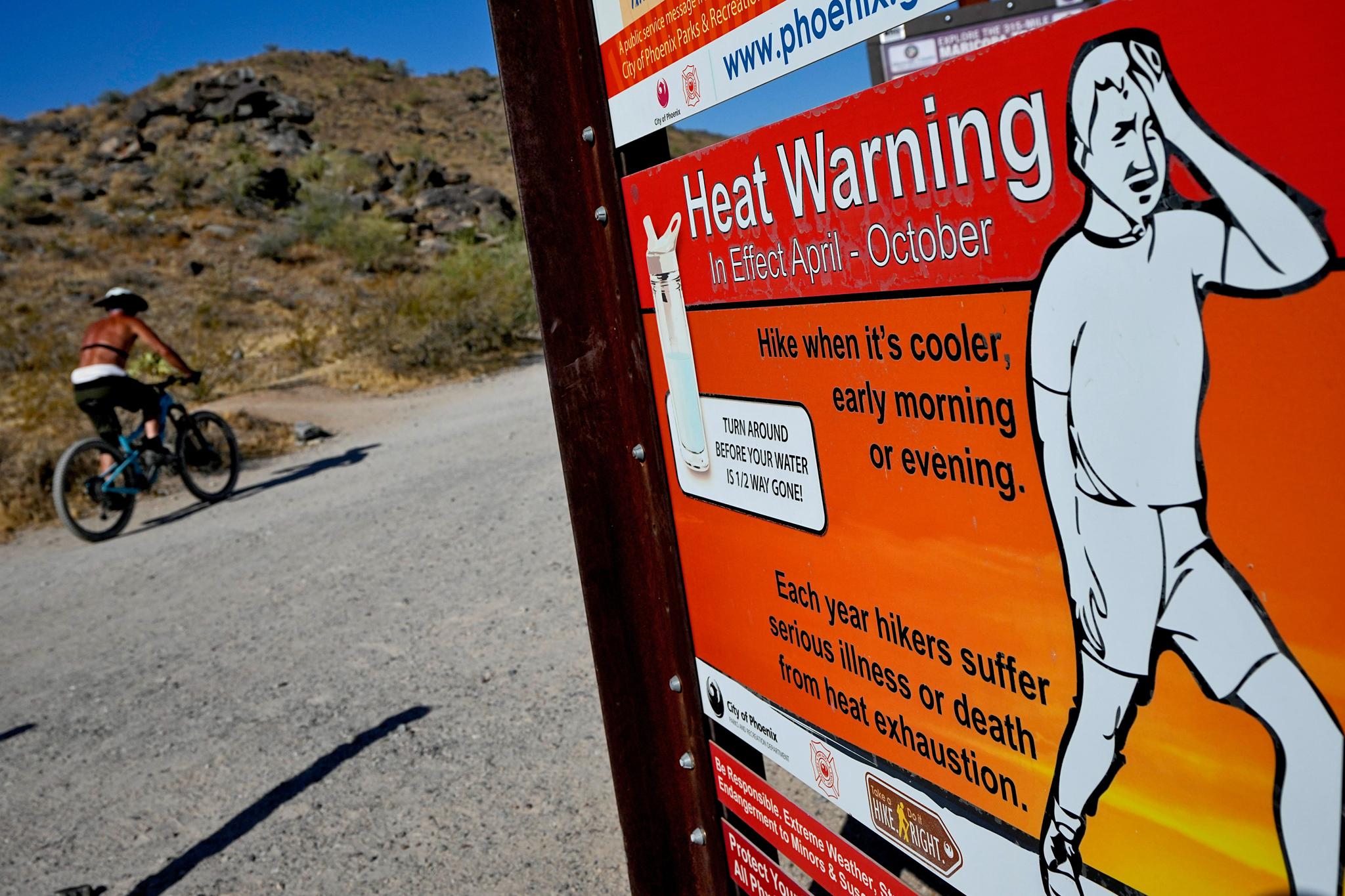 Shirtless man riding uphill next to a heat warning sign