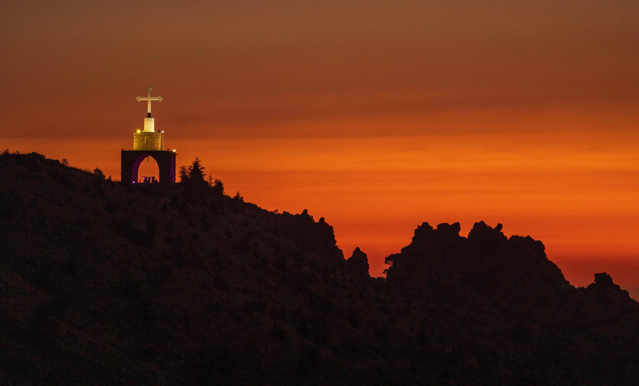An arch on a hill with a cross and an orange/golden sunset behind the mountains