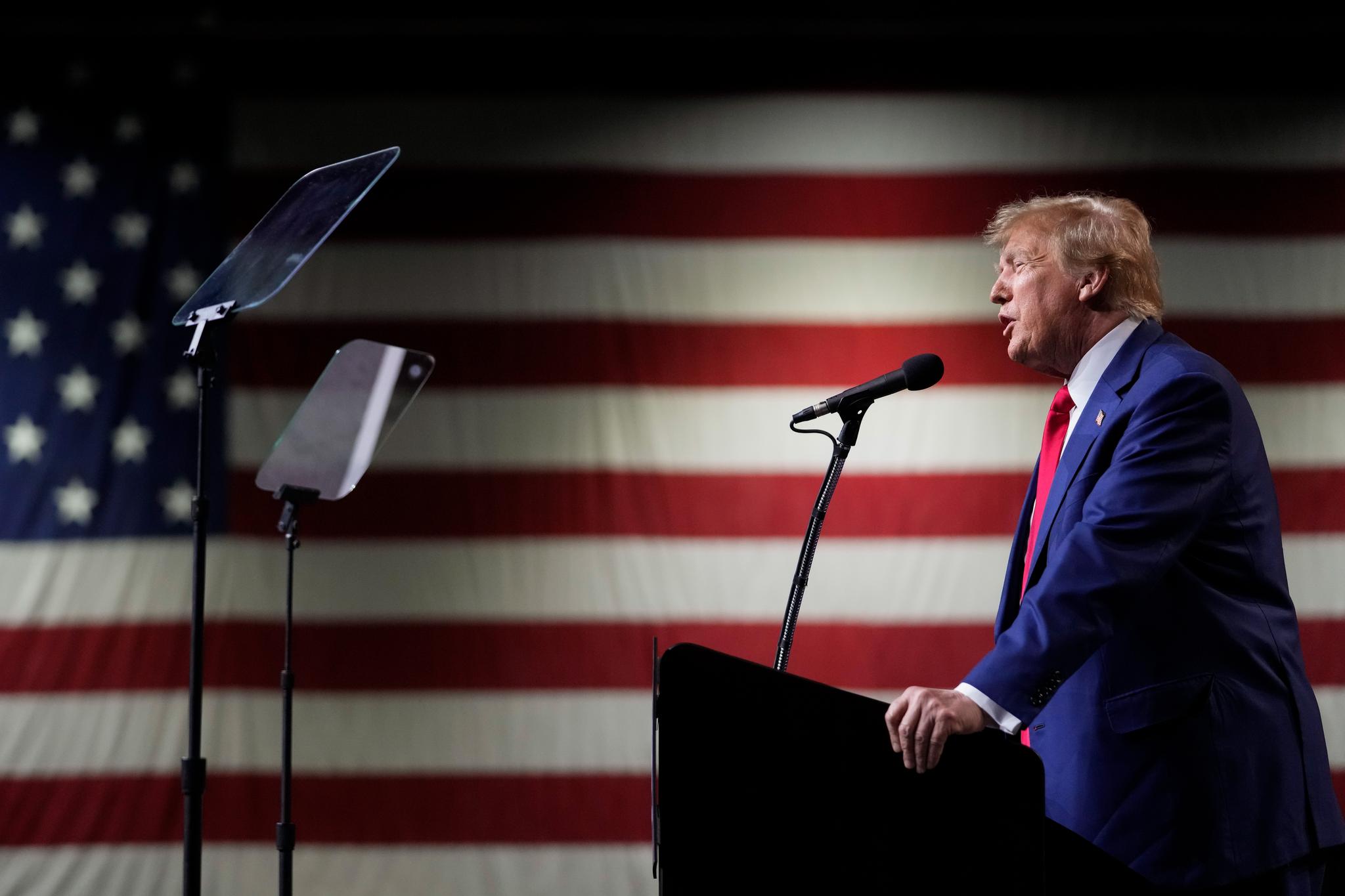 Former Pres. Trump speaking into a microphone with a very large American flag behind him