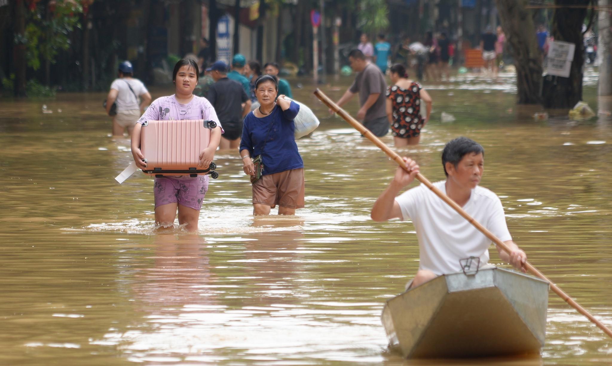 People carrying belongings in knee-deep water and one man rowing a boat in the flood.