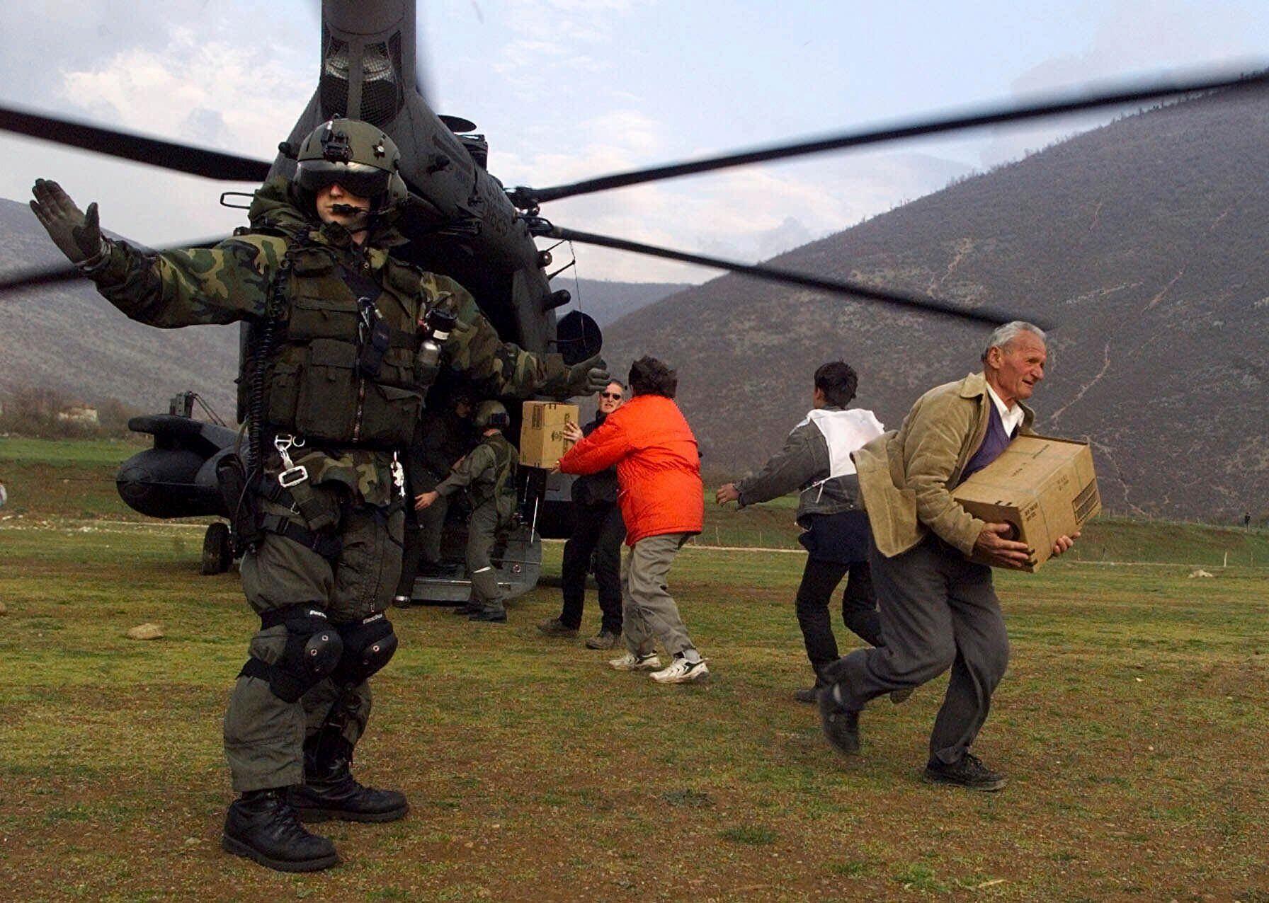 Soldier in front of a helicopter as citizens are unloading boxes of food.