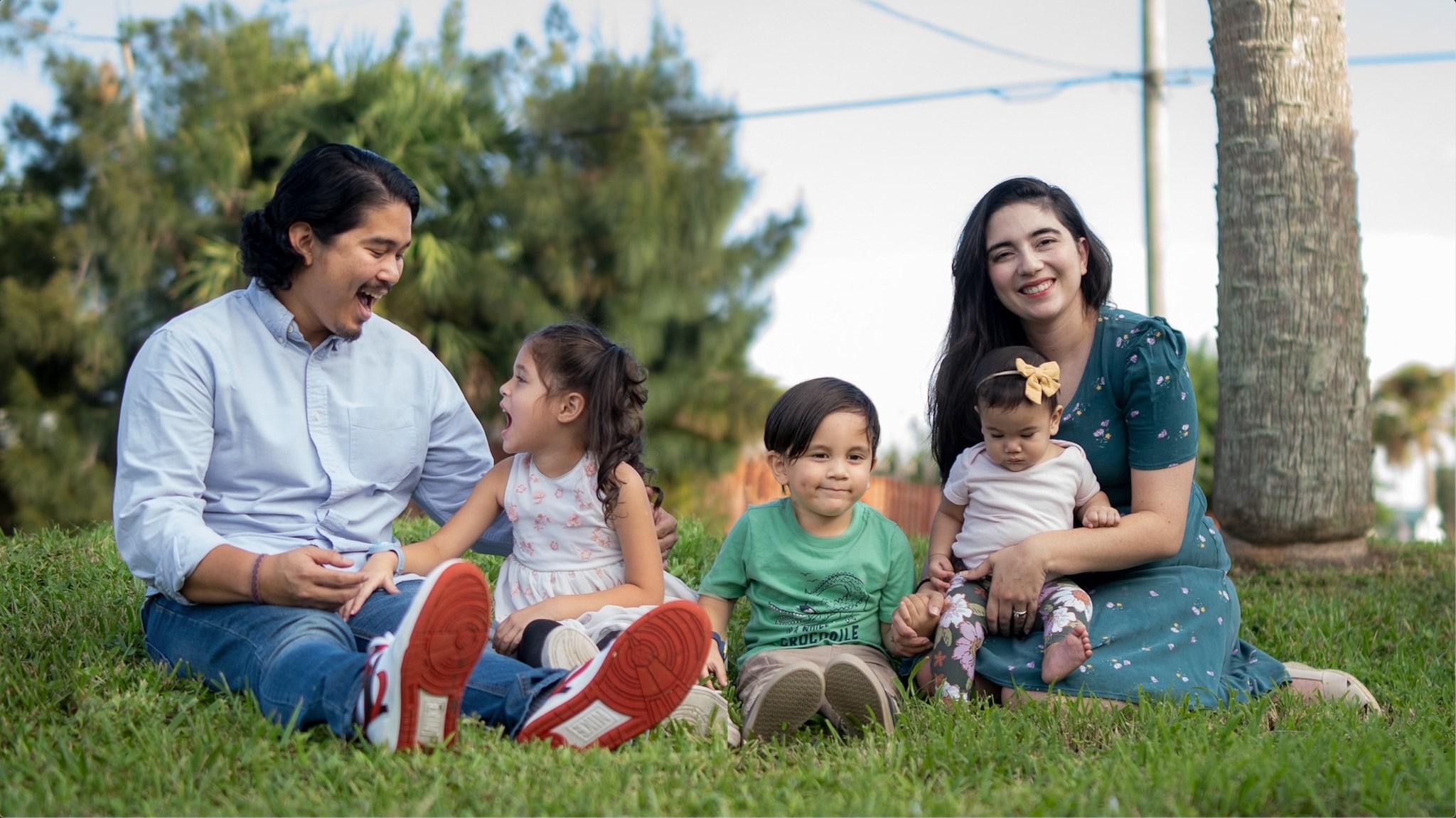 Parents sitting with their three children in the grass, outside, smiling, and laughing as they pose for a family photo. 