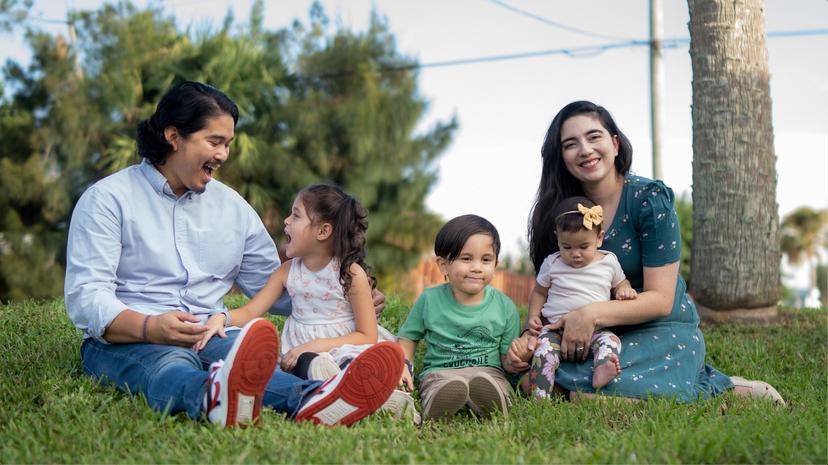 Parents sitting with their three children in the grass, outside, smiling, and laughing as they pose for a family photo. 