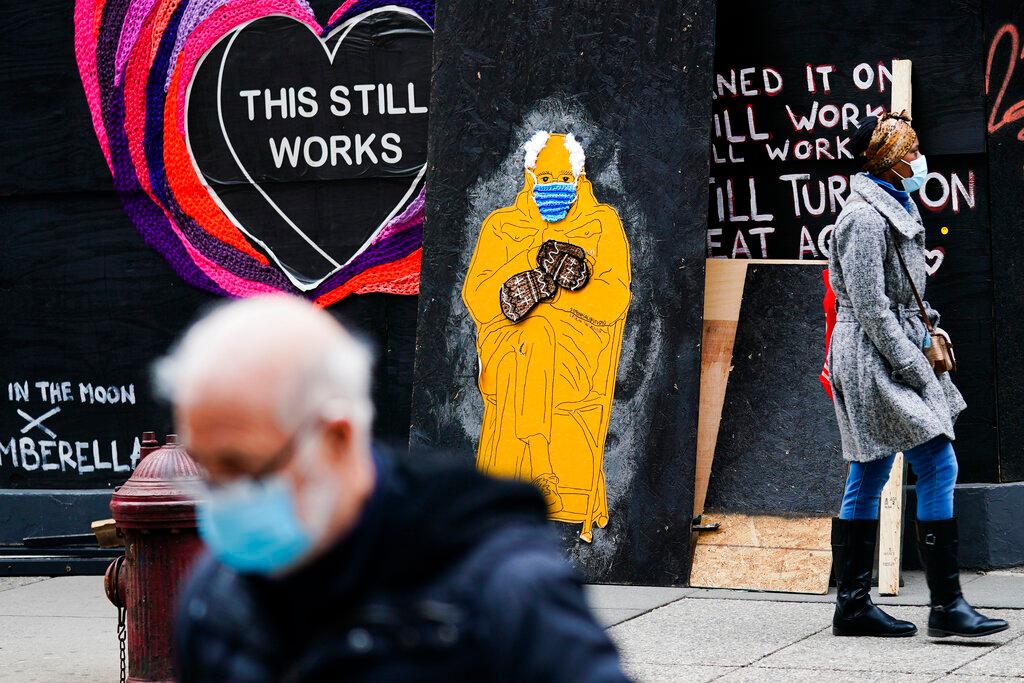 People wearing face masks as a precaution against the coronavirus walk past a depiction of Sen. Bernie Sanders