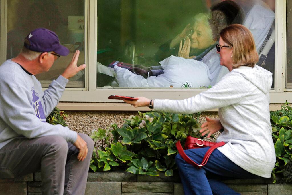 Judie Shape, center, who has tested positive for the coronavirus, blows a kiss to her son-in-law, Michael Spencer, left, as Shape's daughter, Lori Spencer, right, looks on, as they visit on the phone and look at each other through a window at the Life Care Center in Kirkland, Wash.