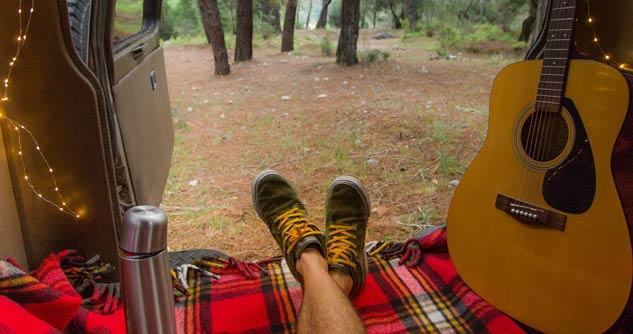 Man sitting in van looking out back at woods, we see his feet, water bottle and his guitar