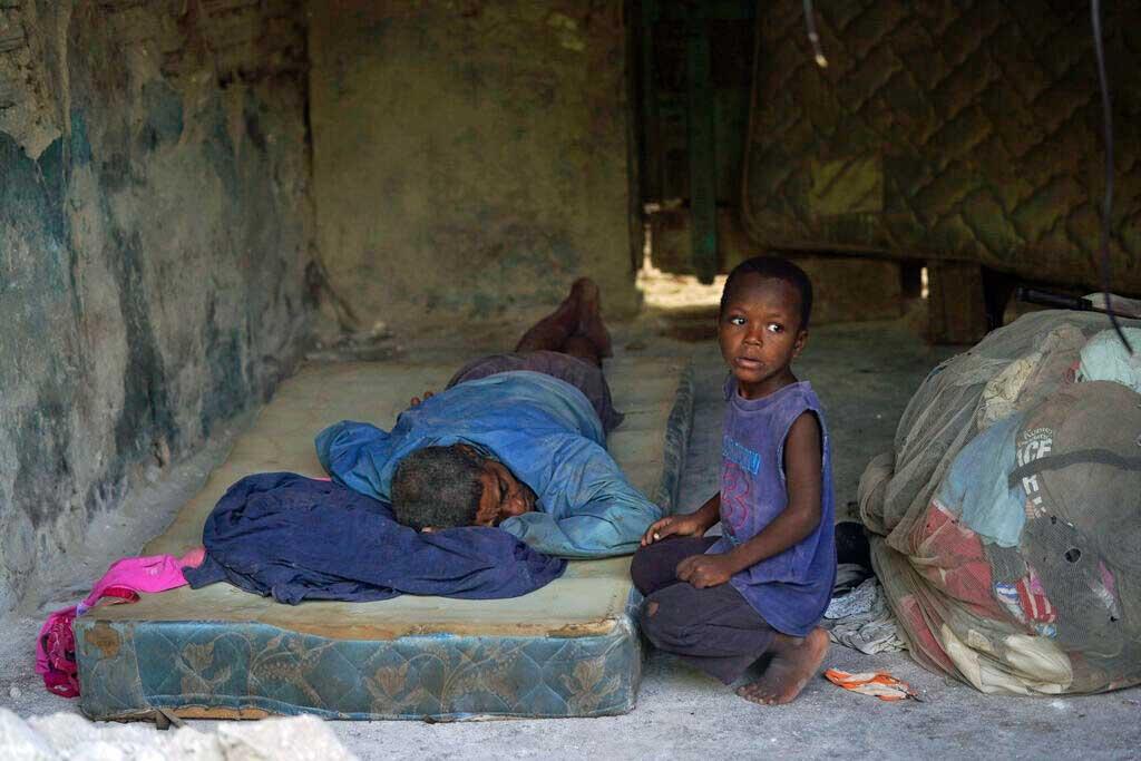 A child crouches next to man sleeping on the porch of his home in Les Cayes, Haiti 