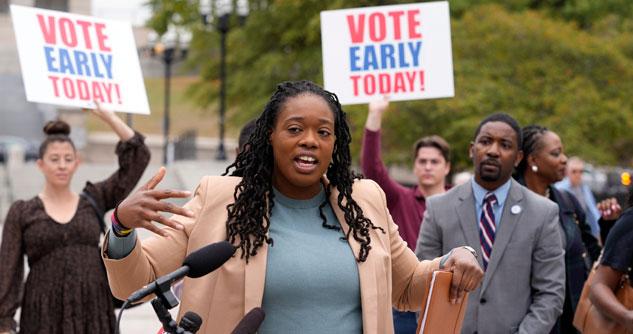 Woman in blue shirt and light pink blazer speaking with people in the background holding signs that say "Vote Early Today"