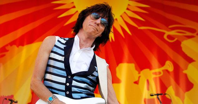 Man with a white guitar and a blue and black striped vest in front of a bright orange and yellow background