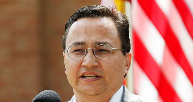 Man with glasses standing in front of a microphone with an American flag in the background