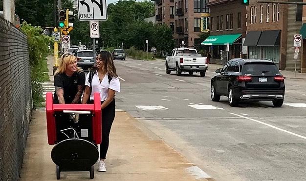 Red salon chair wheeled along sidewalk by two women